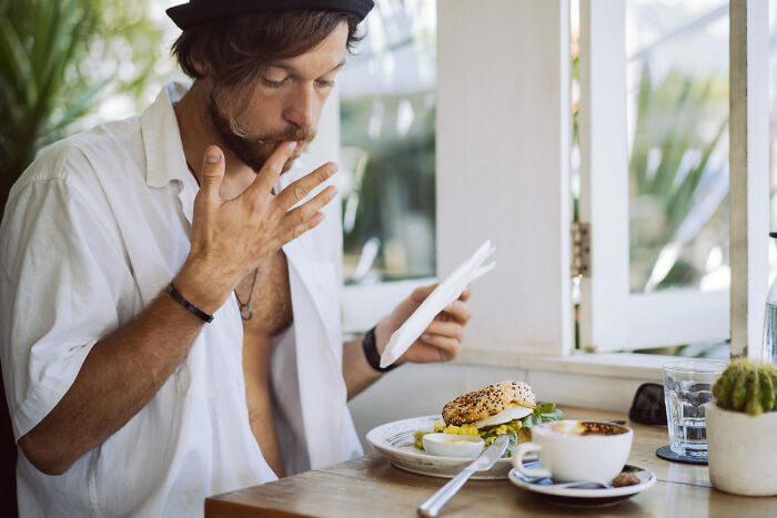 Man eating a bagel sandwich in a cafe, displaying non-physical qualities.