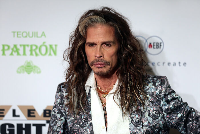 A celebrity with long hair and floral jacket at an event, background features sponsor logos.