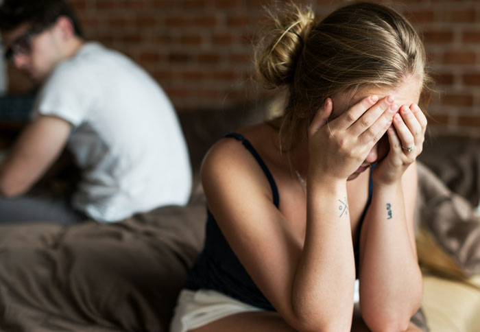 Woman with hands on face, appearing upset, while man sits on bed, facing away.