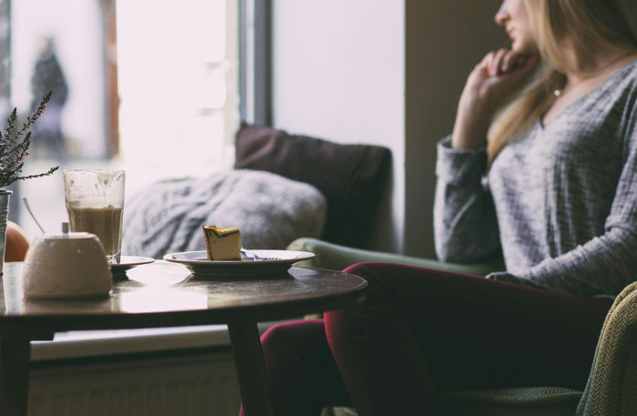 Woman in a light sweater seated at a table in a restaurant, looking thoughtful with dessert and drink nearby.