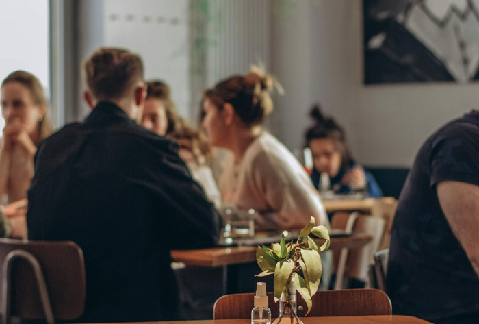 People talking in a café, with a plant in the foreground; setting for discussing uncovering backgrounds.