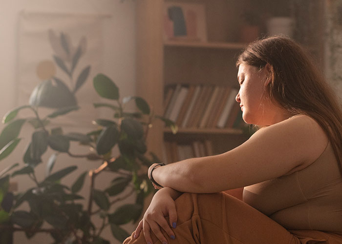 A woman sitting thoughtfully by a plant, reflecting on challenges of being overweight.