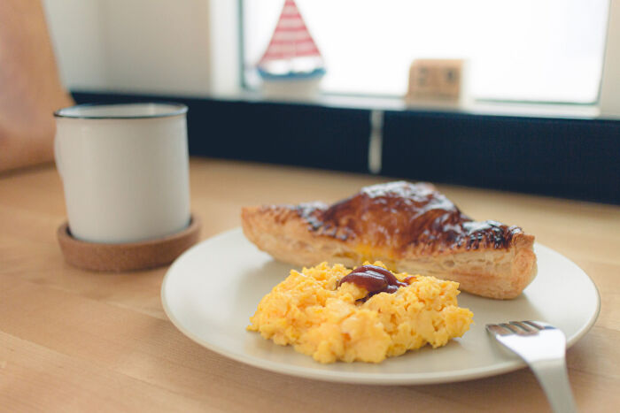 Plate with scrambled eggs and pastry on a wooden table next to a white mug, illustrating dessert experiences and food commonality.