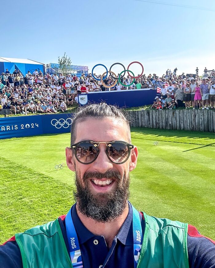 Bearded man taking a selfie in front of a crowd at Paris 2024 Olympic venue, wearing sunglasses and a badge.