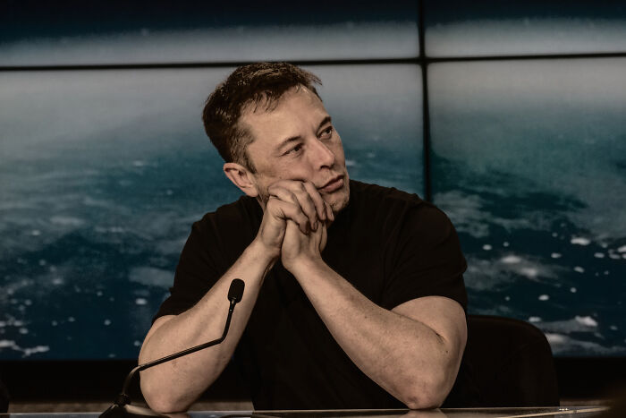 A celebrity sitting at a microphone, pondering, against a backdrop of space imagery.