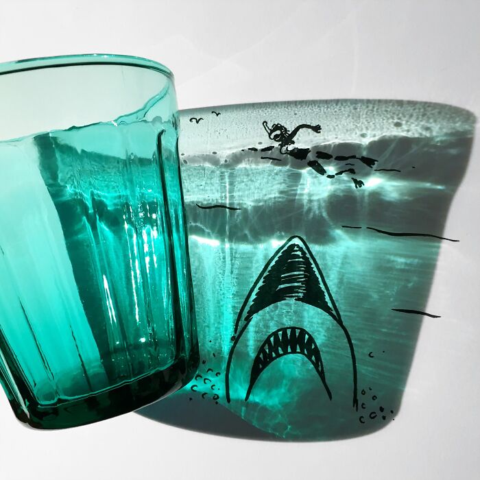 Glass casting a shadow transformed into an illustration of a shark and swimmer, showcasing artist's creative shadow illustrations.