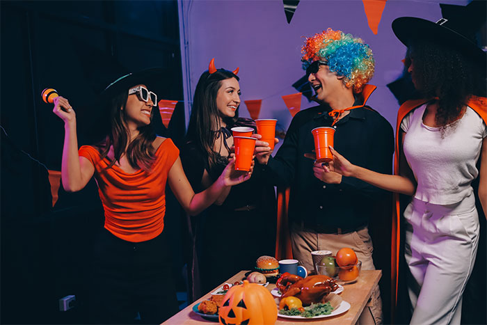 Friends in costume at a Halloween party, laughing and holding drinks, with a decorated table in the foreground.