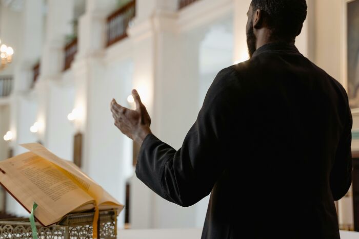 Person speaking in a church, gesturing with hands, beside a large open book.