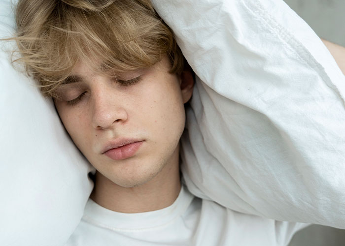 Young man resting with eyes closed, face framed by a white pillow, conveying tranquility and introspective moment.