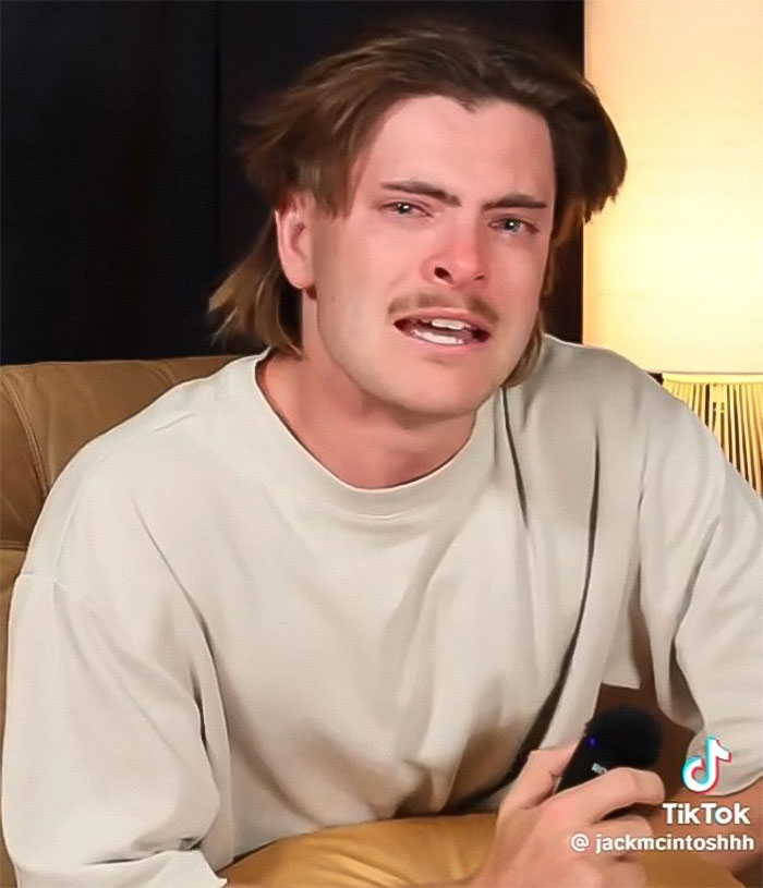 Podcaster in a beige shirt on a couch, visibly emotional, holding a microphone.