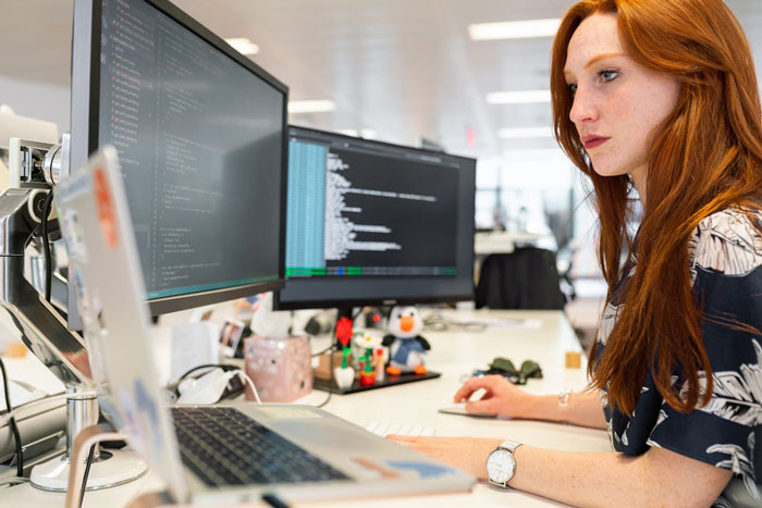 Woman sitting at a desk with multiple monitors, automating her job tasks, coding visible on screens.