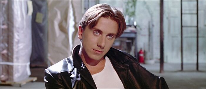 Actor in a leather jacket with a 90s hairstyle, giving a relaxed expression in an industrial setting.
