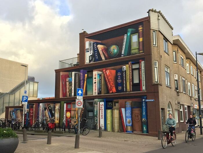 Hyperrealistic mural by Jan Is De Man depicting a bookshelf illusion on a city building wall.