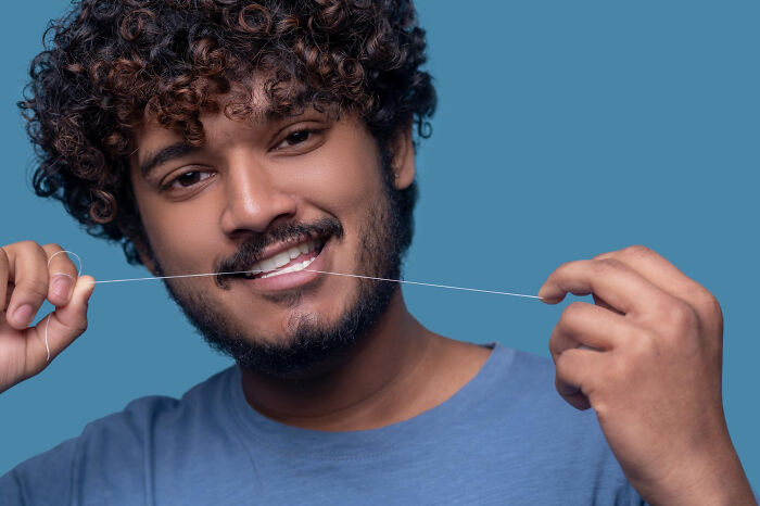 A man with curly hair flossing teeth, promoting common hygiene habits against stinky issues, in a blue shirt on a blue background.