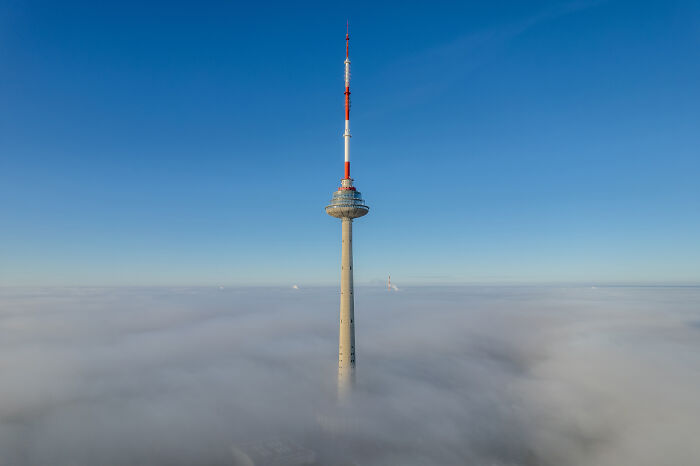 Lithuania's TV tower emerging through clouds, showcasing a stunning aerial view against a clear blue sky.