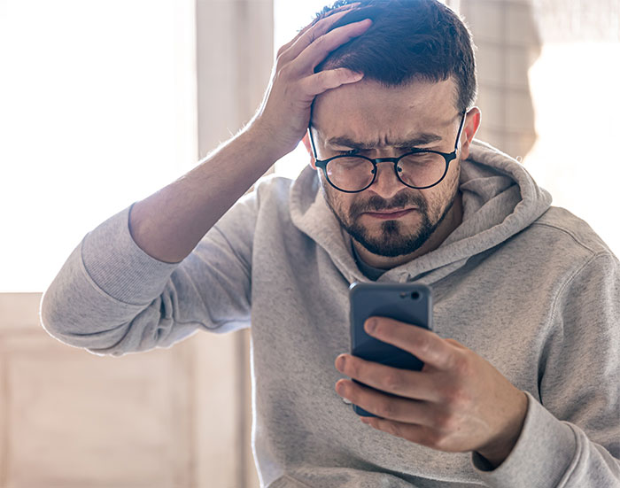 Man in glasses and gray hoodie, looking at phone with confused expression, thinking about system issues. Man in glasses and gray hoodie, looking at phone with confused expression, thinking about system issues.