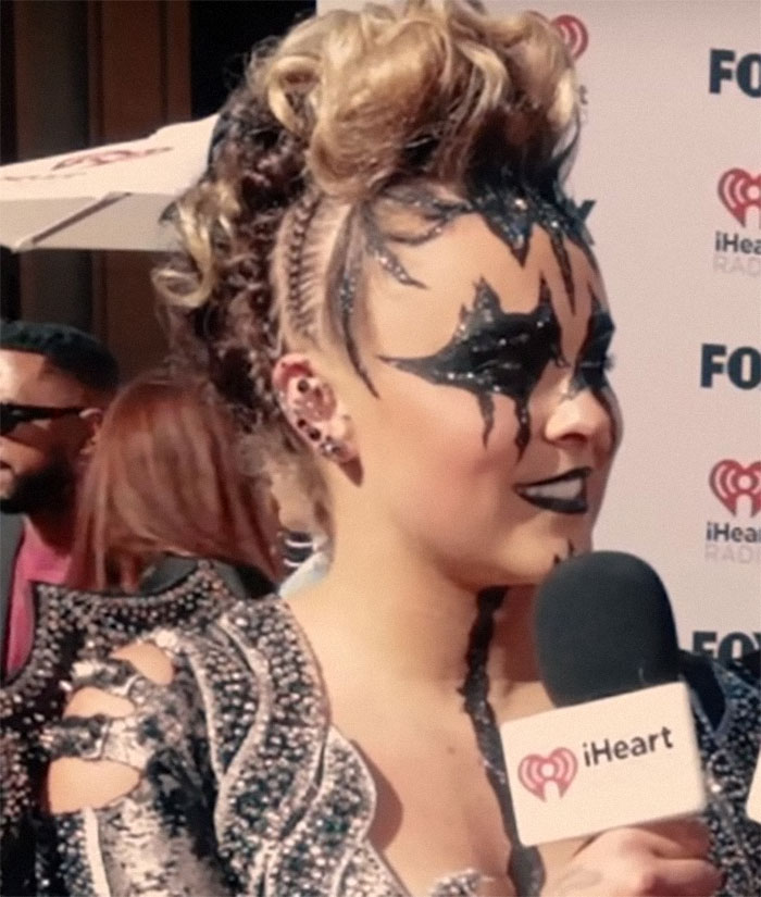 Singer at iHeart event with dramatic makeup and braided hairstyle, speaking into a microphone.