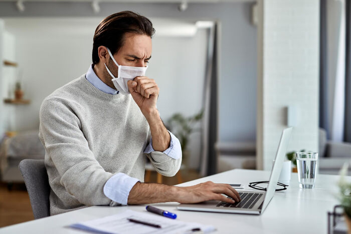 Man wearing mask, coughing at desk while working on a laptop, illustrating American norms during health concerns.