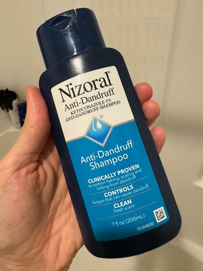 Hand holding Nizoral Anti-Dandruff Shampoo in a bathroom setting.