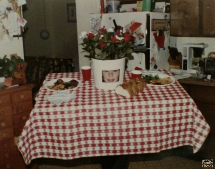 Table with red checkered cloth, a bread loaf, plates of food, and a flower pot with a face cutout, an awkward family photo moment.