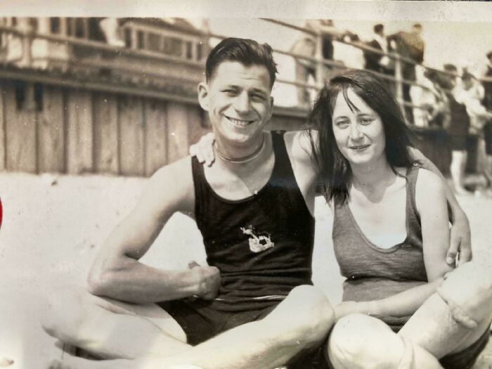 Vintage photo of a smiling couple in swimsuits sitting on a beach, showcasing retro summer fashion and joy.