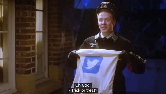 Person in costume holding a bag with a Twitter logo, standing outside a house at night. Subtitled: "Oh God! - Trick or treat?"