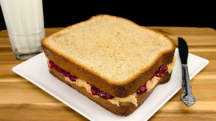 Giant peanut butter and jelly sandwich on a plate with a glass of milk, humorously oversized to evoke nostalgia.