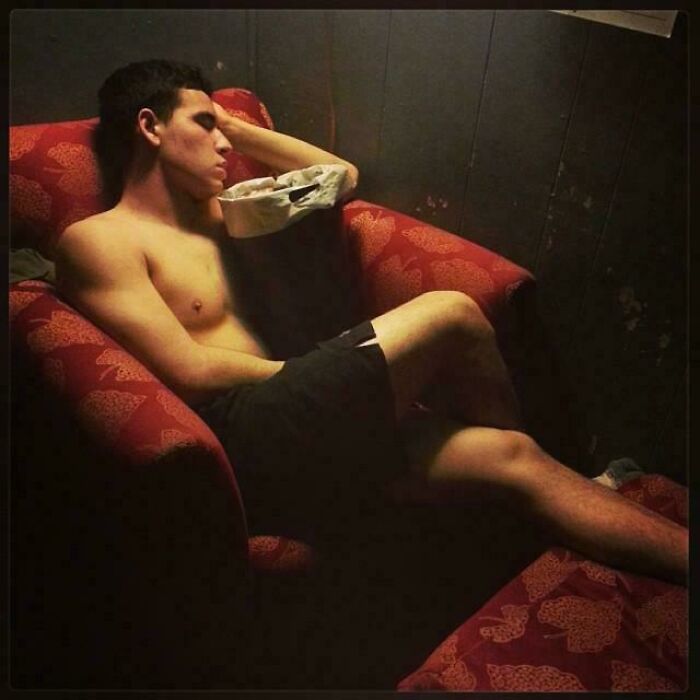 A shirtless man sleeps on a patterned red armchair, evoking an accidental renaissance feel with soft lighting.