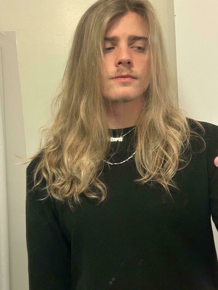 Young man with long blonde hair wearing a black sweater and silver necklaces showcasing fierceflow beautiful hair men style.