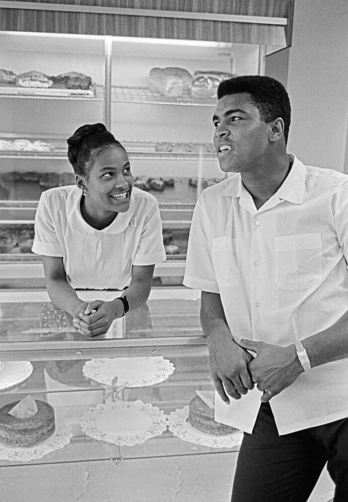 Two people in a bakery, one leaning on the counter, sharing a light moment. Rare historical photo.