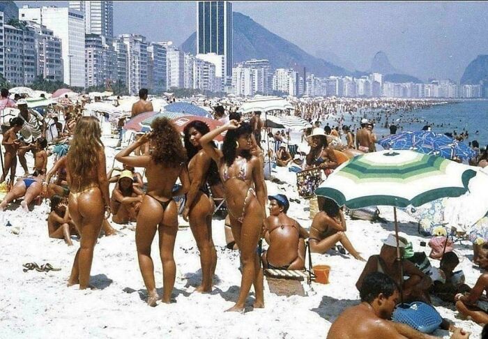 Historical photo of a crowded beach with people sunbathing and umbrellas, city skyline in the background.