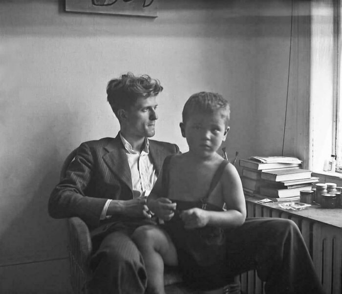"Vintage photo of a man in a suit with a young boy on his lap, seated near a stack of books by a window."