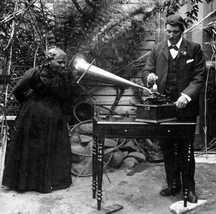 A man operates an early phonograph while a woman in a dress listens, capturing a rare historical moment with vintage technology.