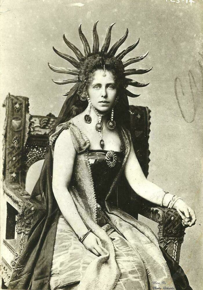 "Historical photo of a woman in regal attire with a unique headdress, seated on an ornate chair."