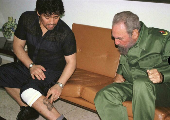 Two men seated on a couch, one showing a leg tattoo to the other, depicting a rare historical moment.