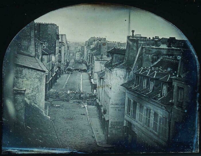 A rare historical photo shows an old cobblestone street bordered by vintage buildings, with a barricade in the distance.