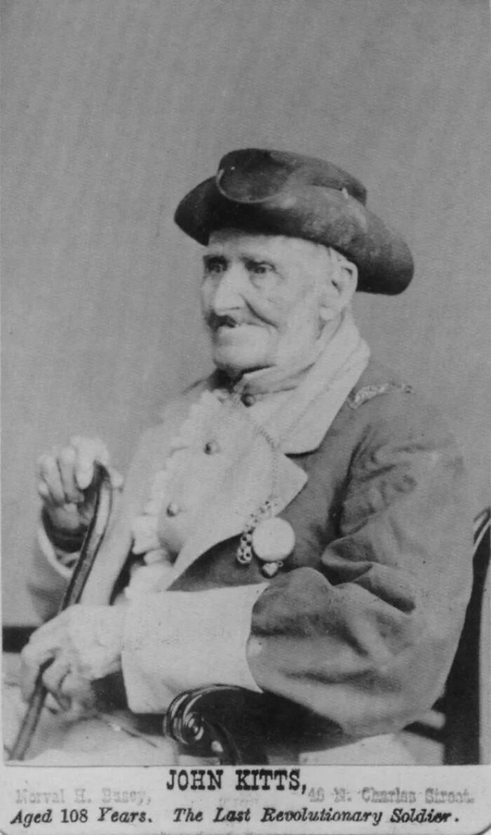 Elderly man in historical attire, identified as John Kitts, seated and holding a cane, representing rare historical photos.