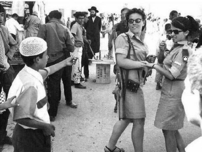 Two women in vintage uniforms interact with a young boy while surrounded by a crowd, depicting rare historical moments.
