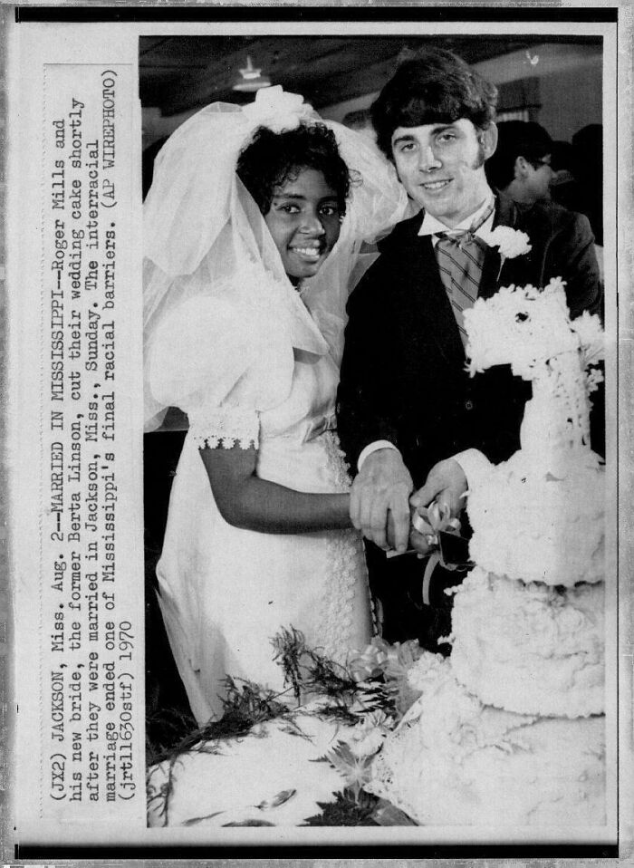 Bride and groom in wedding attire cutting cake in historical photo, symbolizing breaking barriers.
