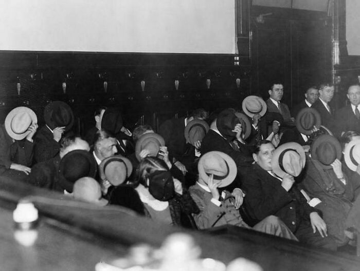 Historical photo showing a group of people, seated indoors, covering their faces with hats.
