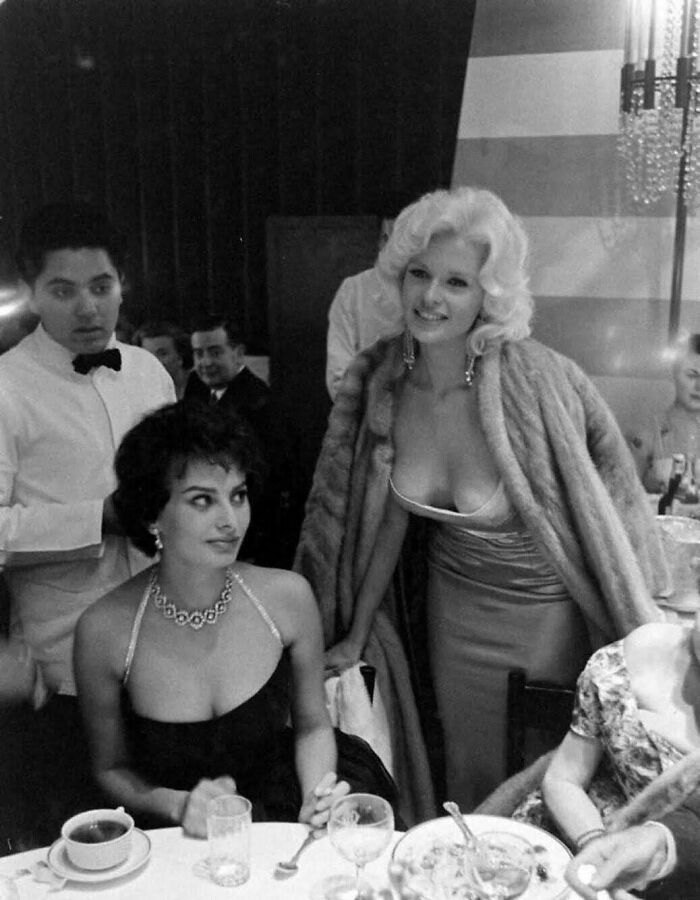 Two women dressed elegantly in a glamorous historical setting, engaging in conversation.