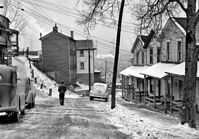 A man walks down a snowy, historical street lined with vintage cars and houses, showcasing rare historical scenery.