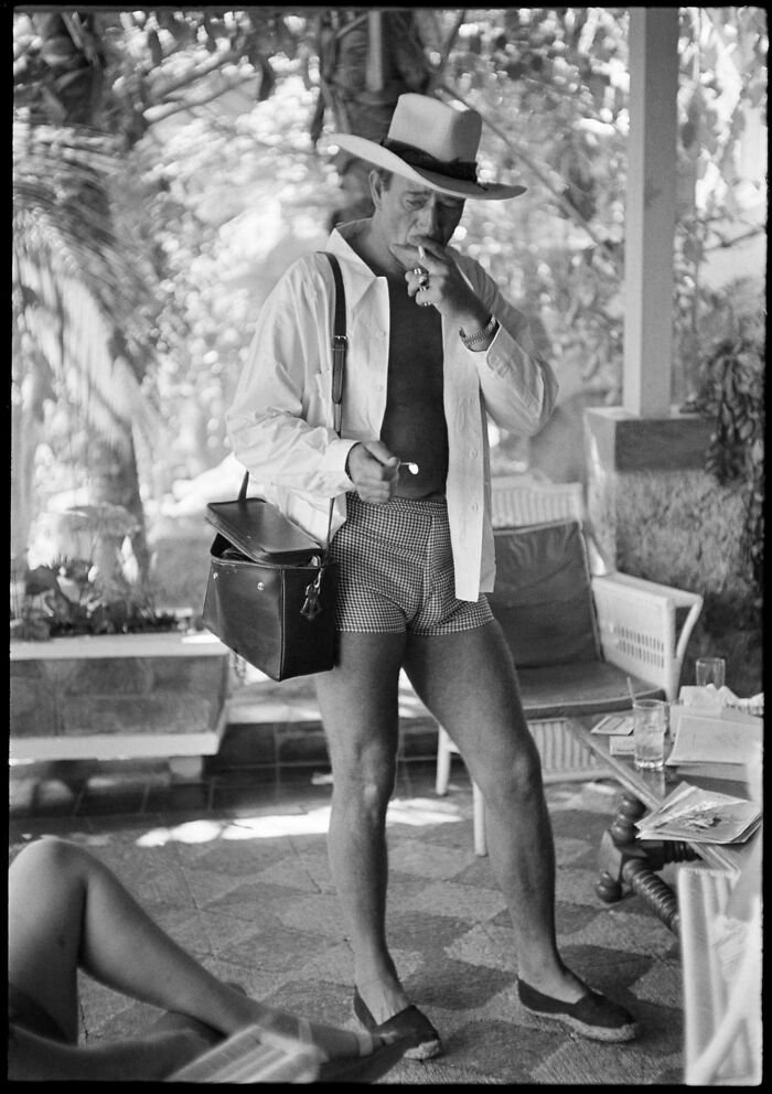Man in shorts and hat smoking, standing on a porch with a satchel. Rare historical photo capturing candid moment.