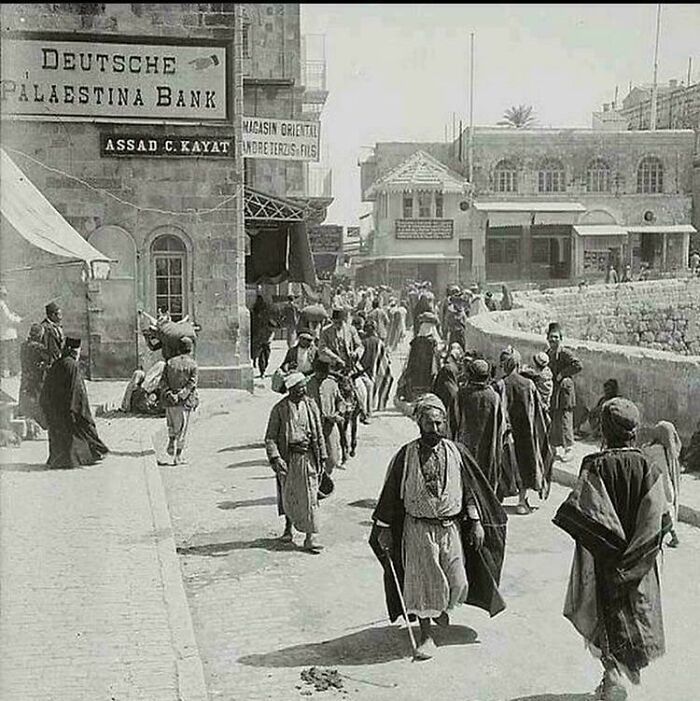 Historic street scene with people walking by a building marked "Deutsche Palästina Bank," reflecting rare historical moments.