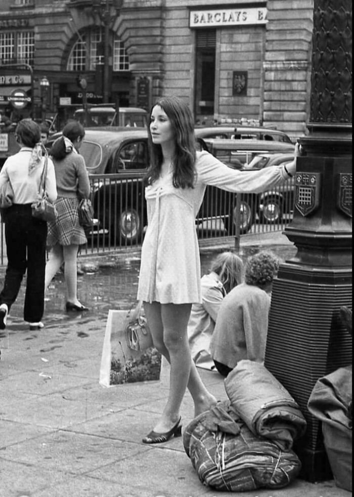 Woman standing on a city street in a white dress, holding a bag; rare historical photo with vintage cars in the background.