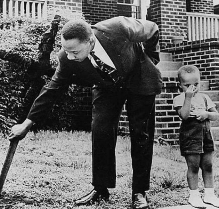 A man in a suit bends down outside a brick building, holding a wooden object near a small child.