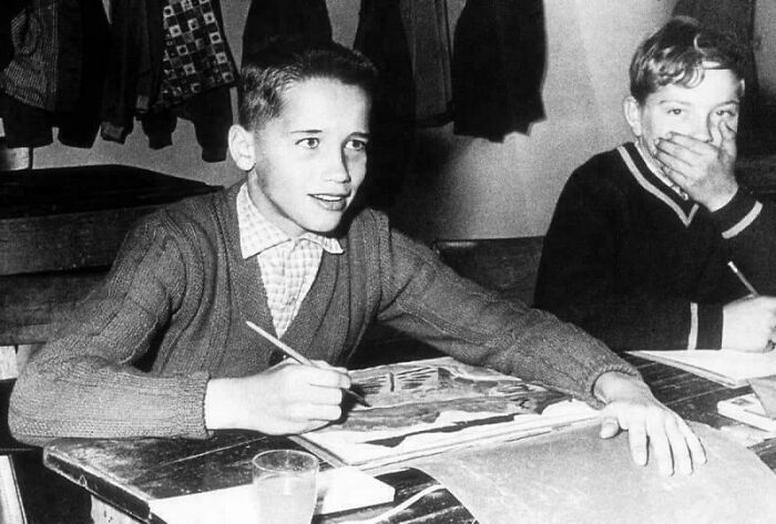 Two boys in a classroom from a rare historical photo, one drawing seriously, the other covering his mouth, jackets hang behind them.