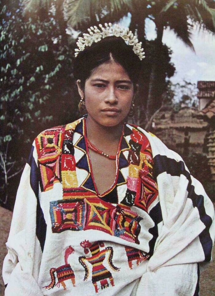 Young woman in traditional embroidered clothing, with floral headpiece, stands outdoors. Rare historical photo.