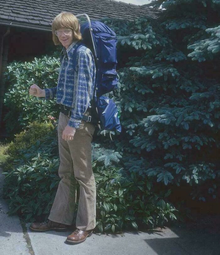 A person with a large backpack, wearing a patterned shirt and bell-bottoms, poses against a backdrop of lush greenery and a roof.