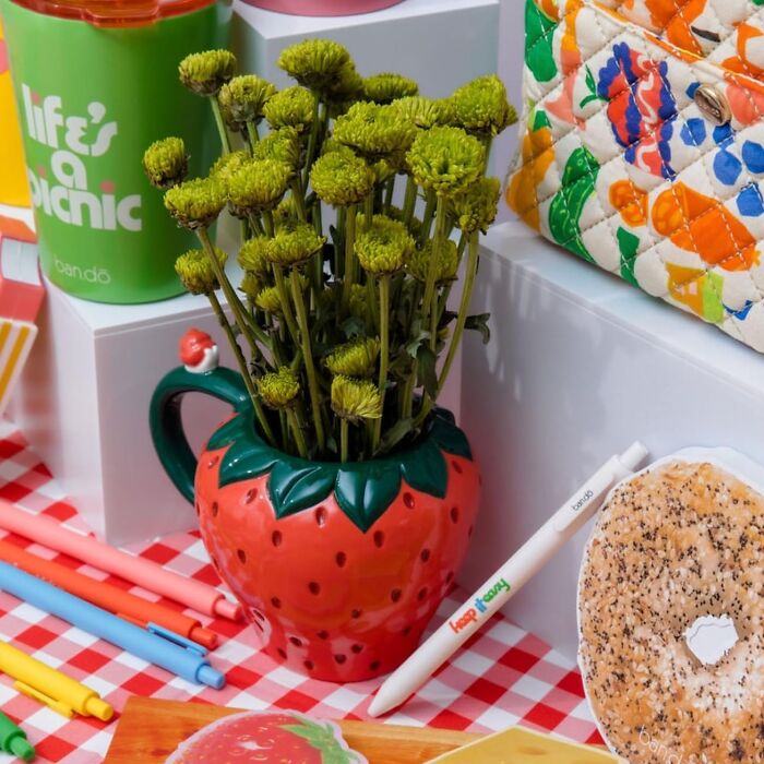 Bando products including a strawberry mug, pens, and a colorful bag on a checkered tablecloth.